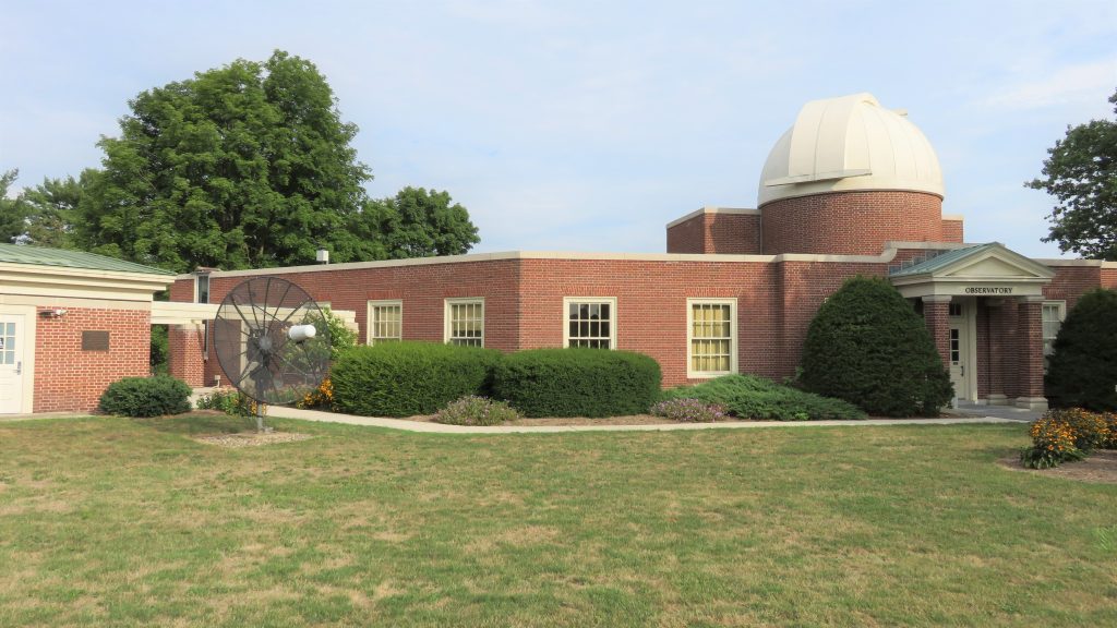 A photograph of the brick observatory building, showing a white dome and a radio telescope dish.